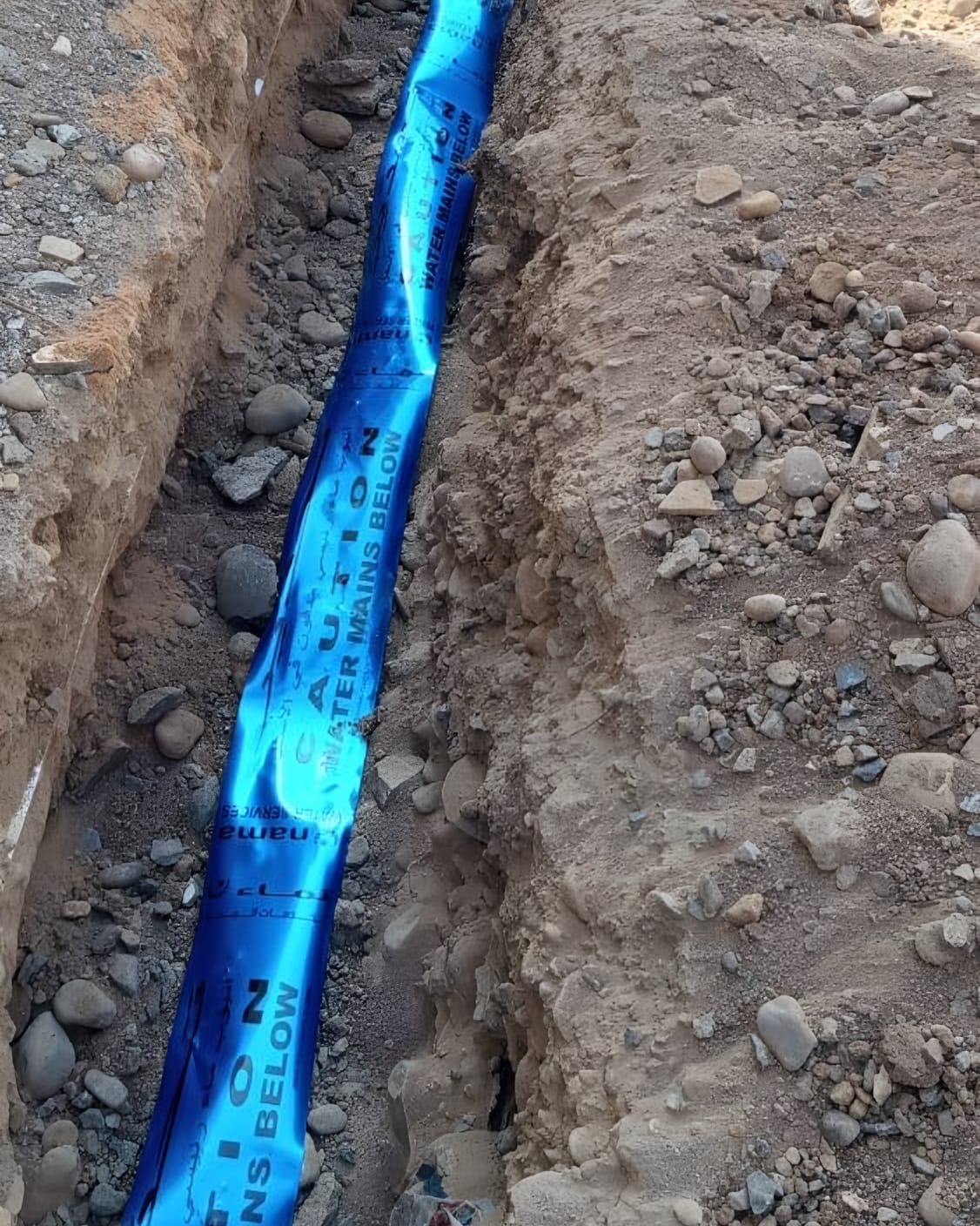 Blue warning tape laid along the trench: “Caution, water mains below”